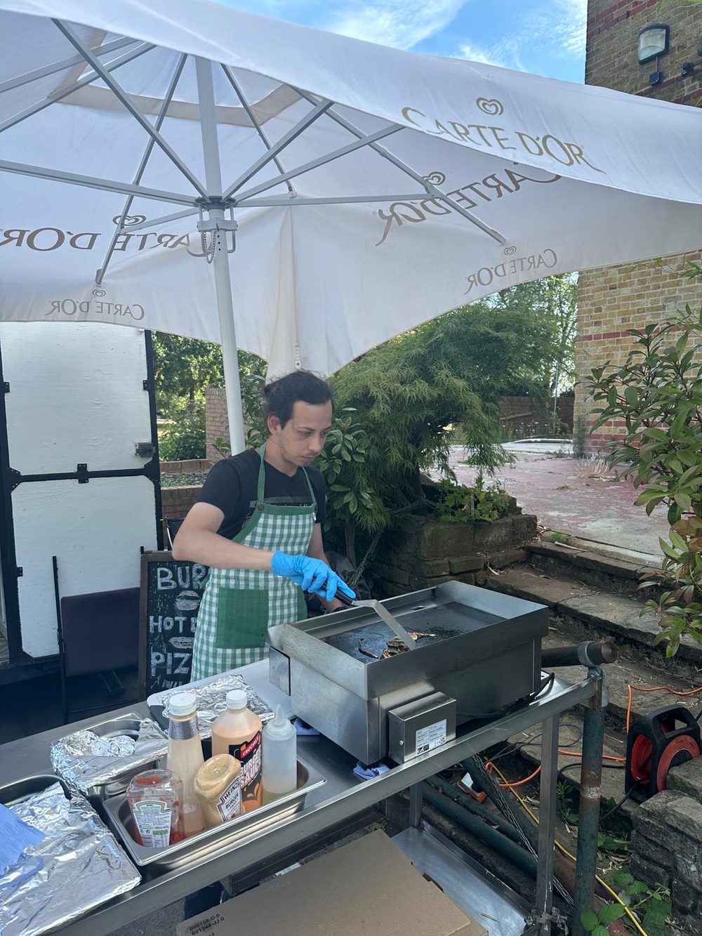 A member of staff in an apron working at the hot-food griddle under a parasol at the outdoor counter