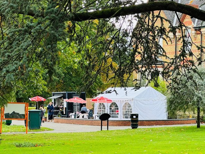 The park lawn seen through overhanging branches, with the marquee, red parasols and playground beyond