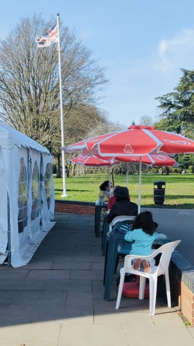 Outdoor paved seating with a red parasol, white chairs and a flagpole, park and sky beyond