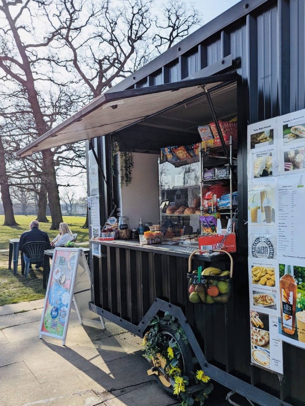 The black cabin-style cafe kiosk in Friary Park with its serving hatch open and fruit and snacks on the counter